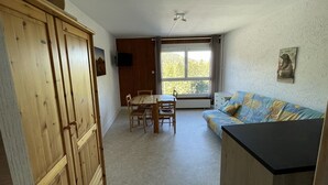 A small, cozy room with a wooden wardrobe, a dining table with chairs, a sofa, and a large window providing a view of greenery outside.