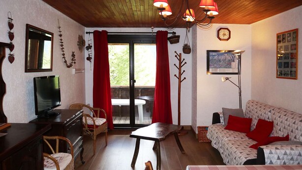 A cozy room with a wooden ceiling, a television, a sofa with red cushions, a dining table, and a large window with red curtains.
