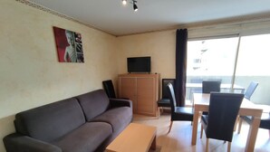 A living room with a sofa, a coffee table, a TV on a cabinet, and a dining area with chairs and a table. - 3-room apartment in Isola 2000 center, parking, near slopes (Isola 2000)