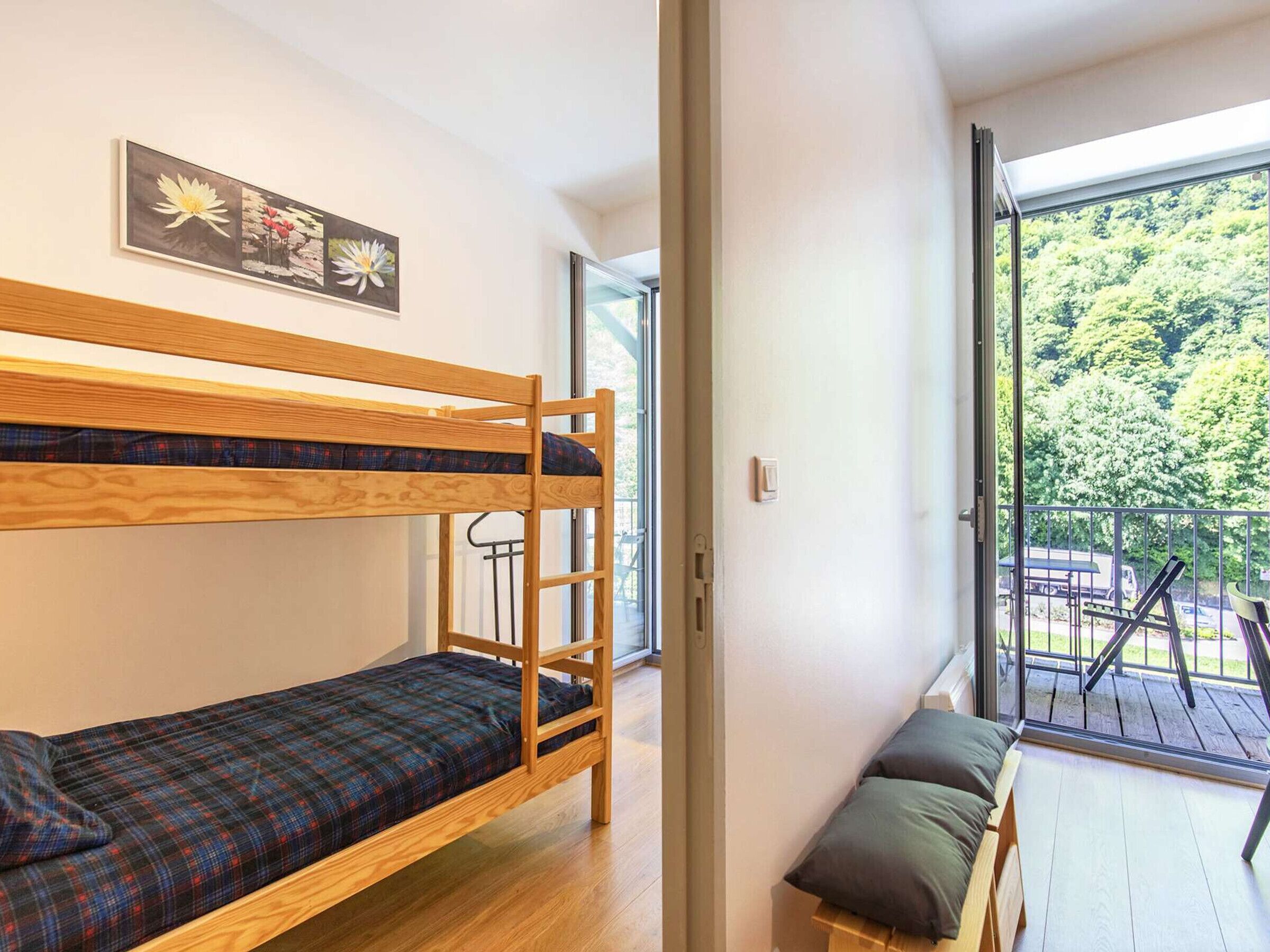 A bunk bed with a plaid bedspread, a small bench with cushions, and a balcony with a view of trees.