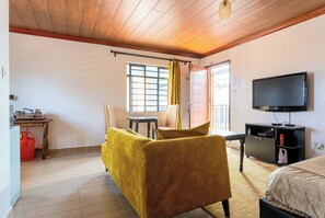 A room with a wooden ceiling, a sofa, a TV, a table, a chair, and a window with curtains.