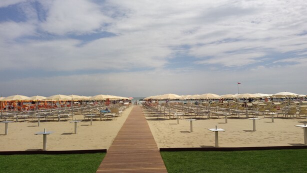 Een strand met rijen ligbedden en parasols, een houten loofpad en een heldere lucht.