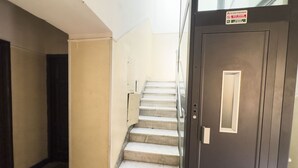 Een trap met een leuning die uitkomt op een plateau, een deur met een bordje, en een eenvoudige muur.