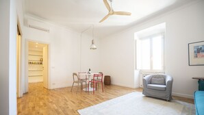 Een ruime kamer met houten vloeren, een plafondventilator, een eettafel met stoelen, een grijze fauteuil en een groot raam dat voor natuurlijk licht zorgt.