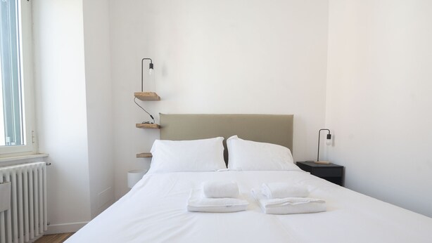 Een net opgemaakt bed met witte lakens en twee opgevouwen handdoeken, een nachtkastje met een lamp, en een radiator op de vloer.