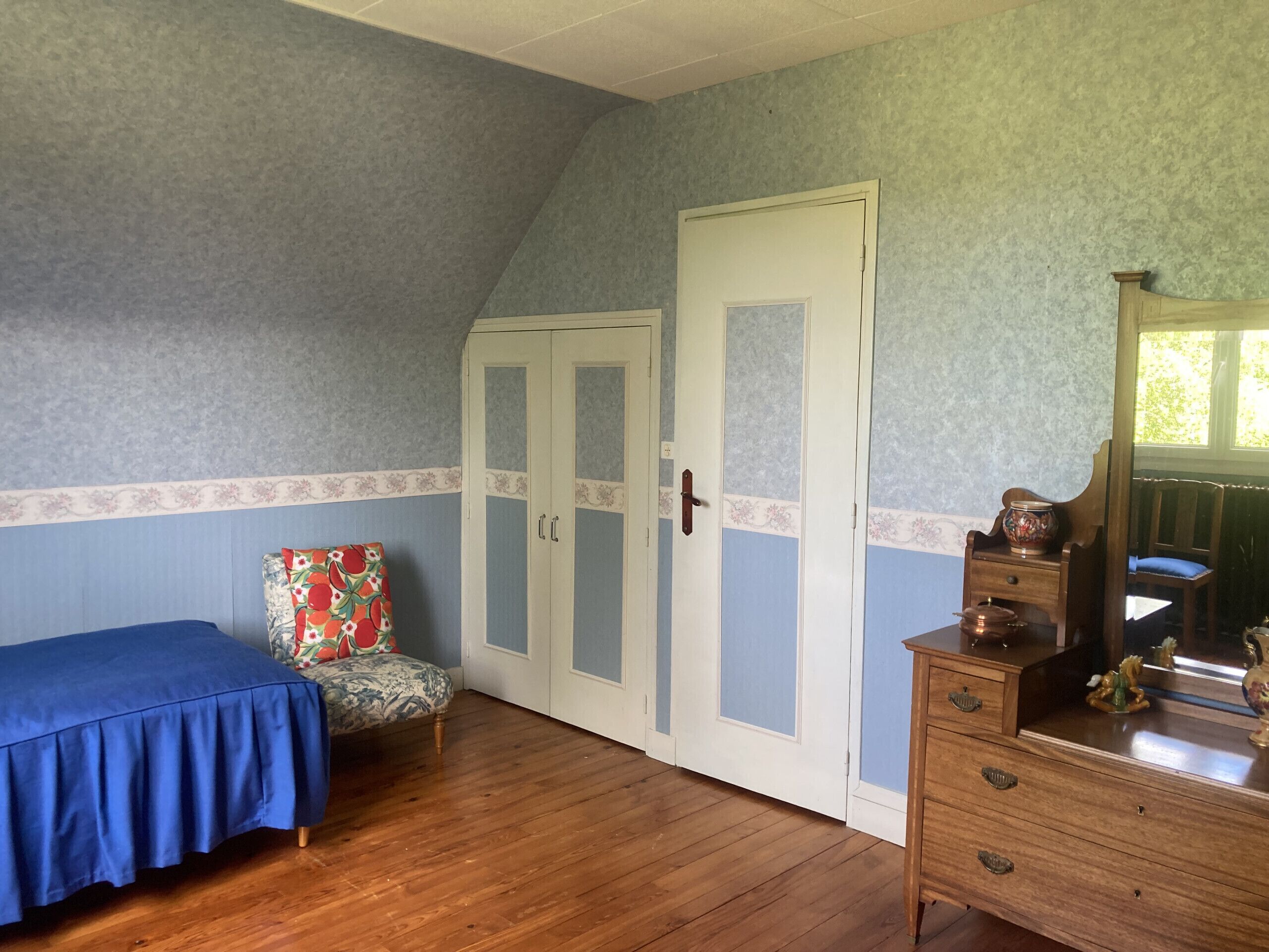Un dormitorio con piso de madera, una cama con cubrecama azul, una silla con cojín estampado, un armario con puertas de cristal y una cómoda con espejo.