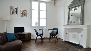 A living room with a brown sofa, two blue chairs, a small round table with a white tablecloth, a TV on a wooden stand, and a fireplace with a mirror above it. - Large studio apartment in the heart of Angers - 2 people (Angers)