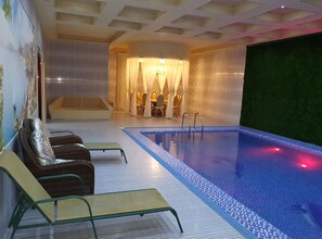 An indoor swimming pool with clear blue water, a lounge area with chairs, and a large screen wall. - Gumbaz Eco SPA Hotel (Samarkand)