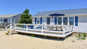 Une maison de plage avec une terrasse en bois, des chaises bleues et une table de pique-nique.