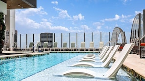 A rooftop pool with lounge chairs, a lifeguard tower, and modern buildings in the background.