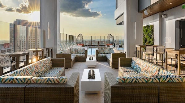 A rooftop lounge with wicker furniture, a central fire feature, and a view of the city skyline.