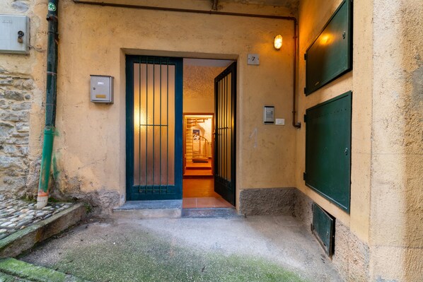 Un estrecho callejón que conduce a la entrada de un edificio, con una puerta verde, un escalón pequeño y suelo de baldosas.