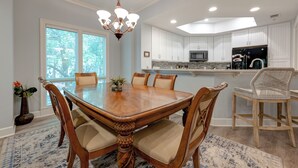 Un comedor con mesa y sillas de madera, una araña y vista a la cocina al fondo.