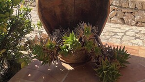 Un vaso rustico e consumato dal tempo con un uccello appollaiato, posizionato su un patio piastrellato circondato dal verde e da un muro di pietra.