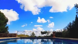 Una piscina con acqua cristallina blu, circondata da alberi e un muro di pietra, sotto un cielo azzurro brillante con nuvole sparse.