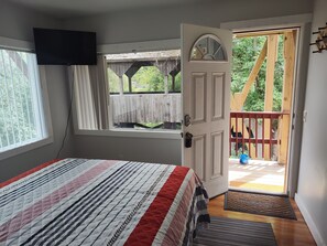 Une chambre avec un lit, un téléviseur fixé au mur, une fenêtre avec des rideaux et une porte ouverte donnant sur un balcon doté d’une terrasse en bois et offrant une vue sur la verdure.
