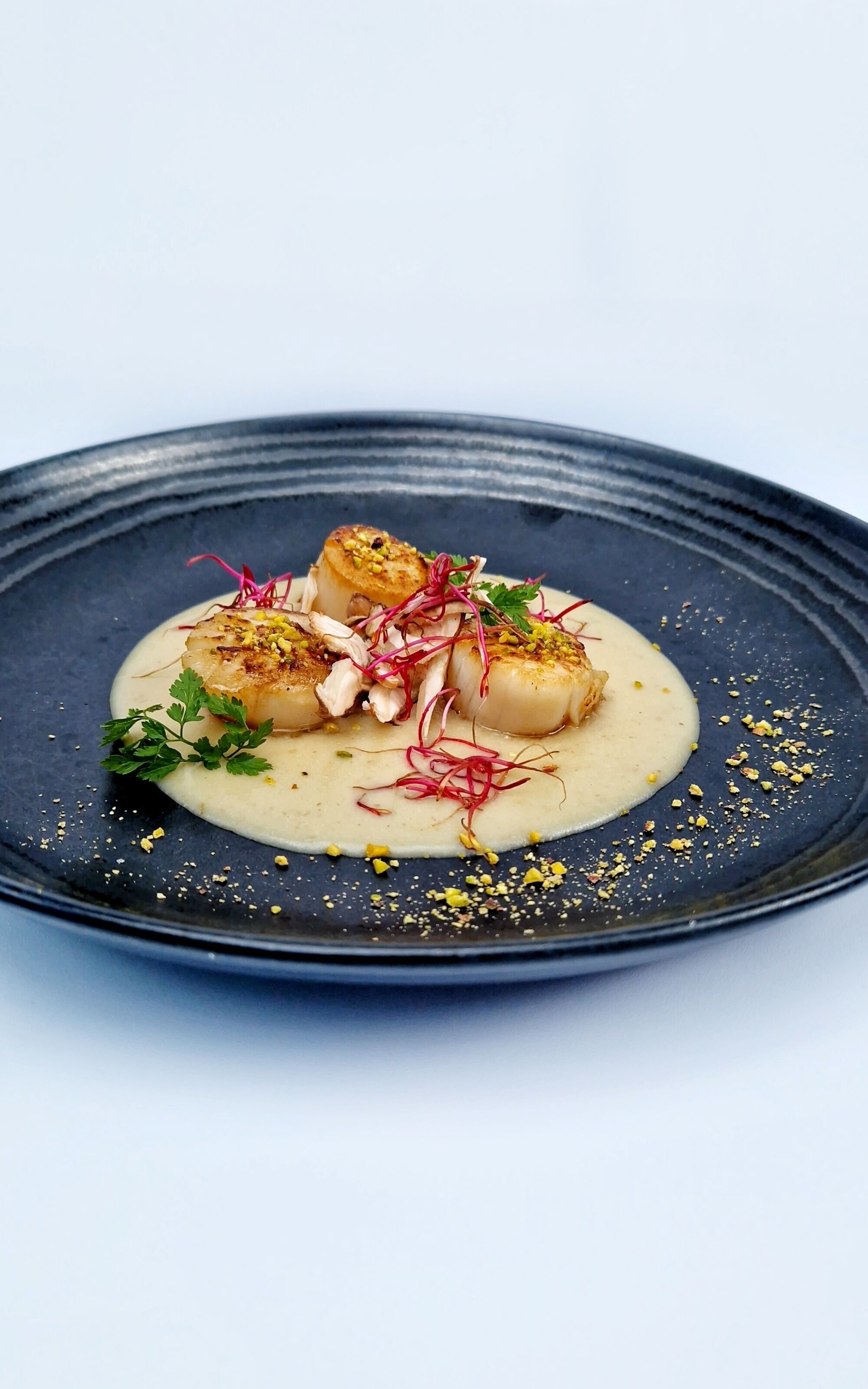 A plate with a dish of seared scallops, garnished with herbs and microgreens, on a dark plate.