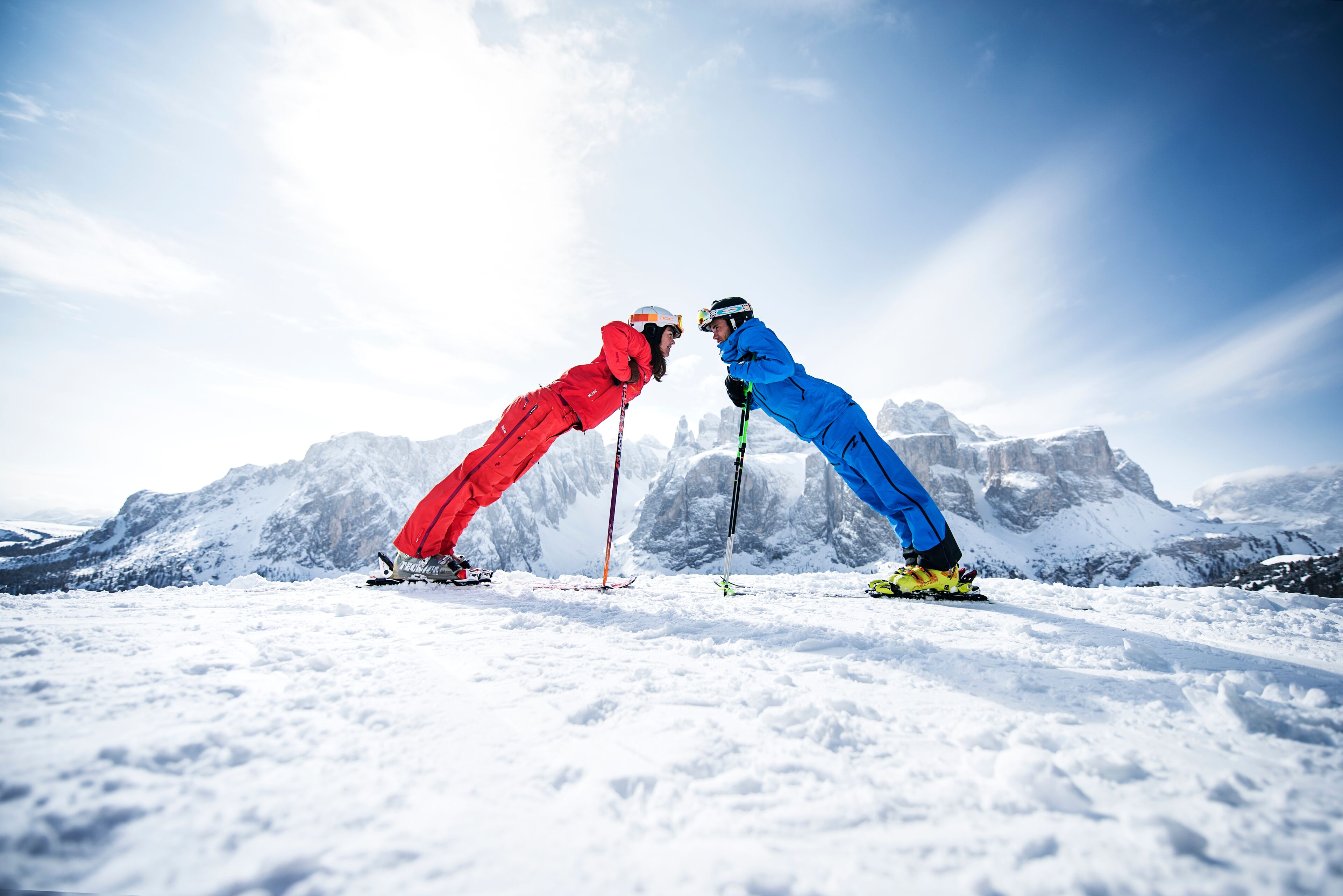 Two skiers in red and blue gear facing each other on a snowy mountain with a clear sky.