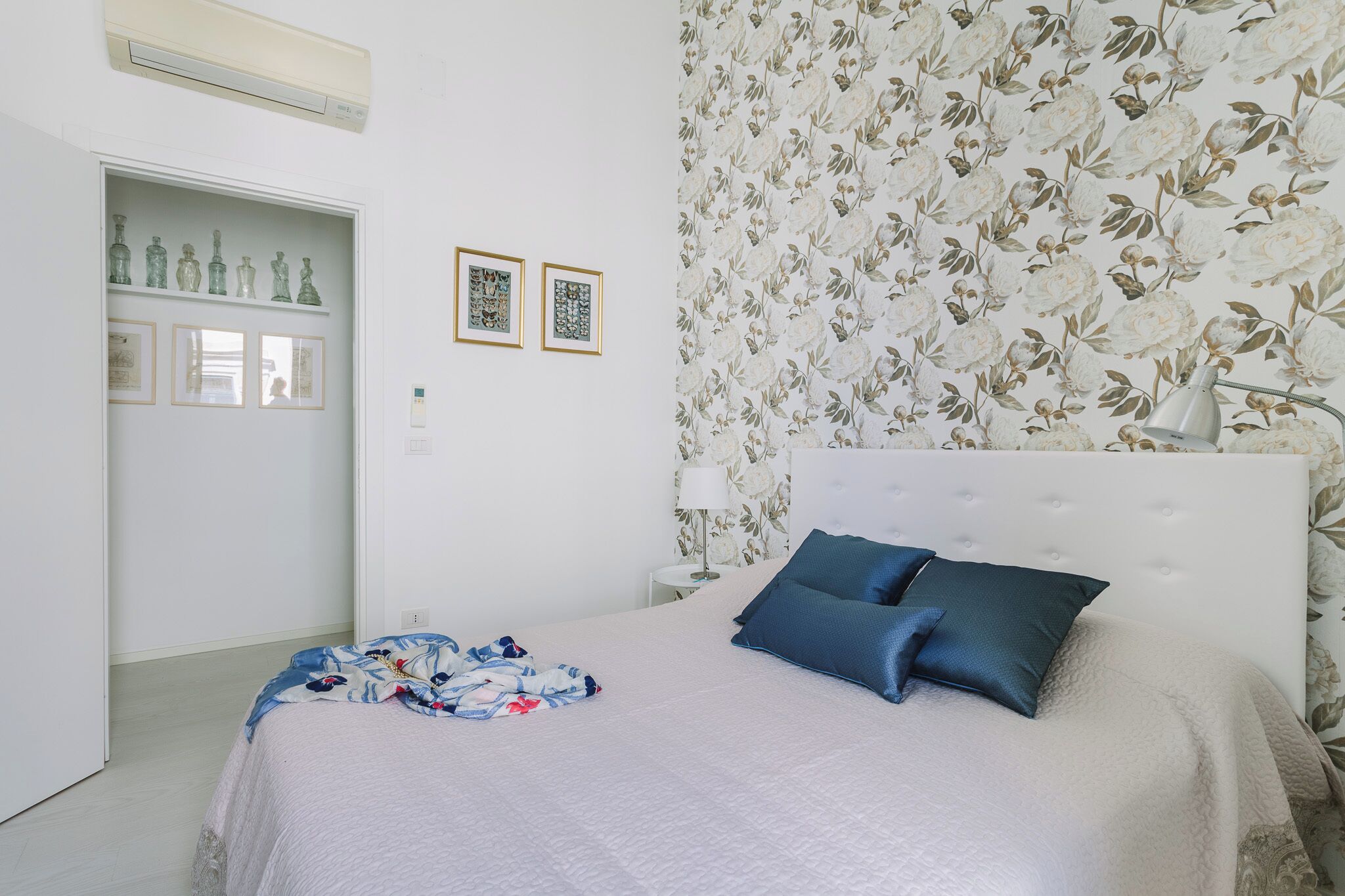 A bedroom with a bed, pillows, a nightstand, a lamp, and a floral wallpaper.