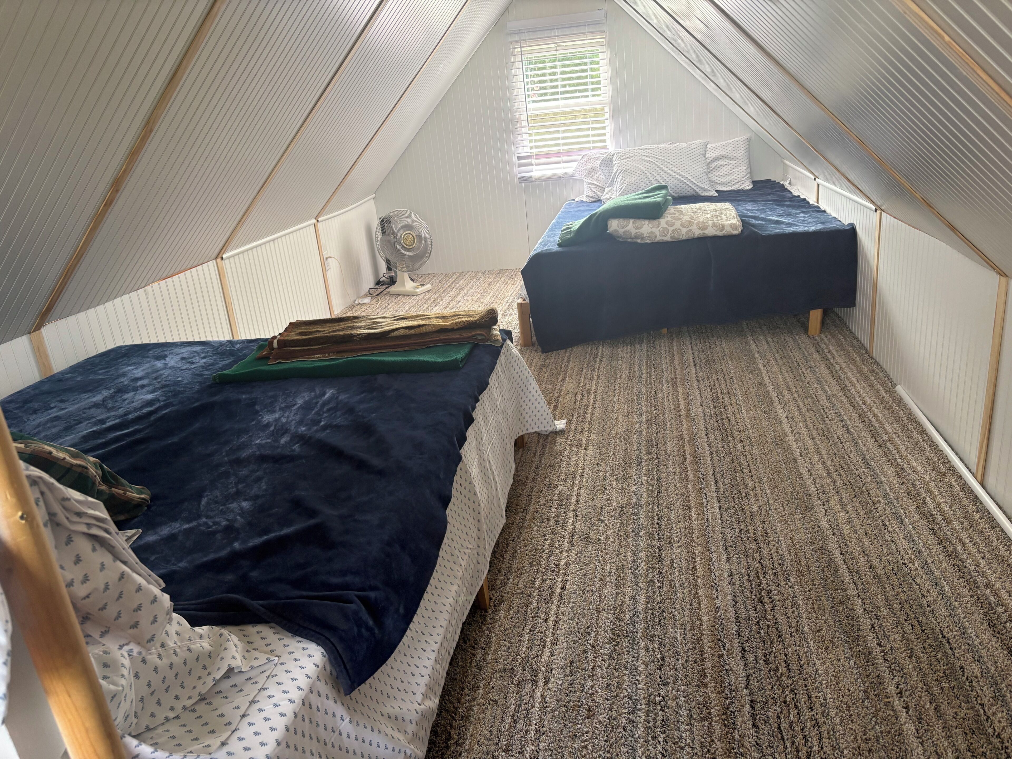 A small attic bedroom with two beds, a fan, and a window.