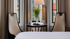 Una habitación de hotel con cama, dos sillas, una mesita con un libro y un jarrón de flores, y una ventana con cortinas.