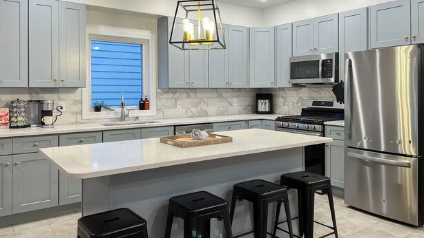 Une cuisine moderne avec des meubles gris, un grand îlot central doté de tabourets, un réfrigérateur en acier inoxydable et un luminaire suspendu.