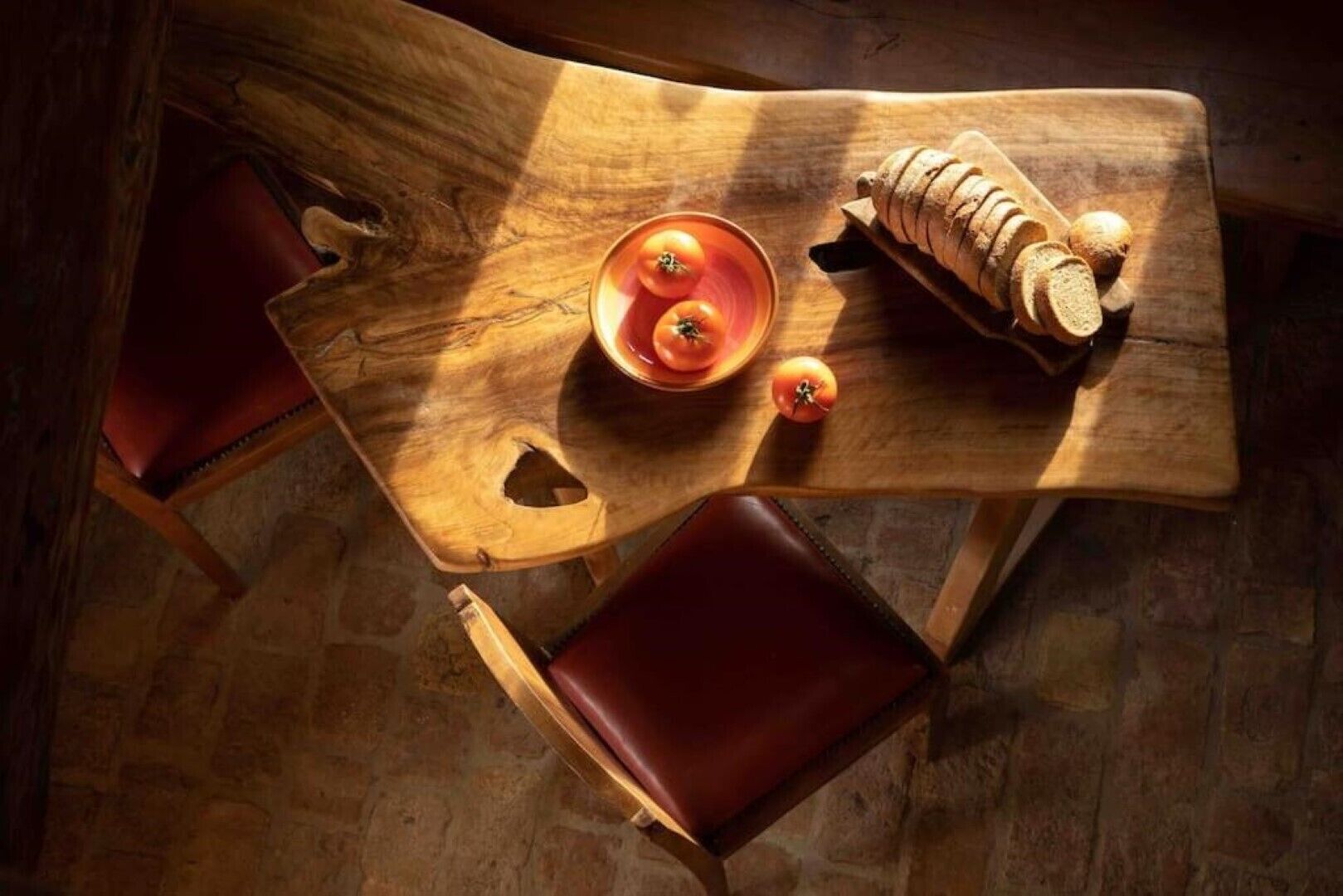 Een houten tafel met een schaal tomaten en sneetjes brood, een stoel en een rustieke stenen vloer.