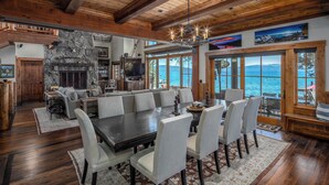 A spacious living area with a large wooden dining table, comfortable seating, a stone fireplace, and a view of the ocean through large windows. - Peaceful Waters by Hauserman Rentals (Tahoma)
