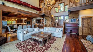 A spacious living area with a large sectional sofa, a wooden coffee table, and a red patterned rug. A piano is visible in the background, and the room features exposed wooden beams and a high ceiling. - Peaceful Waters by Hauserman Rentals (Tahoma)