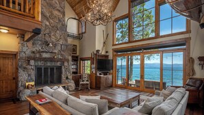 A spacious living room with a stone fireplace, large windows overlooking a body of water, and a cozy seating area. - Peaceful Waters by Hauserman Rentals (Tahoma)