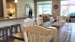 A dining area with a wooden table and chairs, a kitchen in the background, and a large window with blinds. - Sandy Cove Cottage - Steps from the beach #70 (Osoyoos)