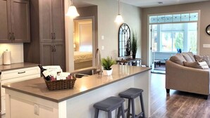 A modern kitchen with dark cabinets, a central island with bar stools, pendant lighting, and a view to a living area with a sofa and a window. - Sandy Cove Cottage - Steps from the beach #70 (Osoyoos)