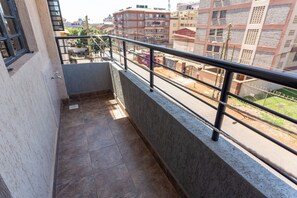 A balcony with a metal railing, a tiled floor, and a view of a cityscape with buildings and a green area.