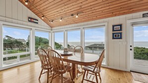 Comedor con mesa y sillas de madera, ventanas amplias con vistas al océano y techo de madera.