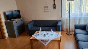 Una sala de estar con un sofá azul oscuro, una mesa de centro de madera con un paño decorativo, un televisor sobre un mueble y una lámpara de pie.