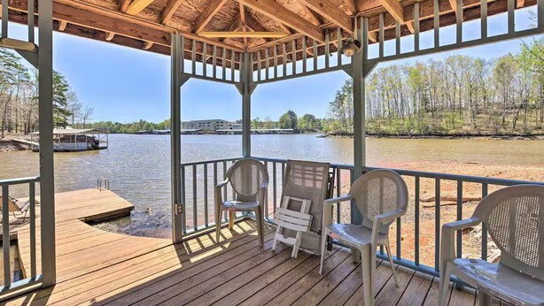 A covered deck with chairs overlooking a body of water, a dock, and a building in the background.