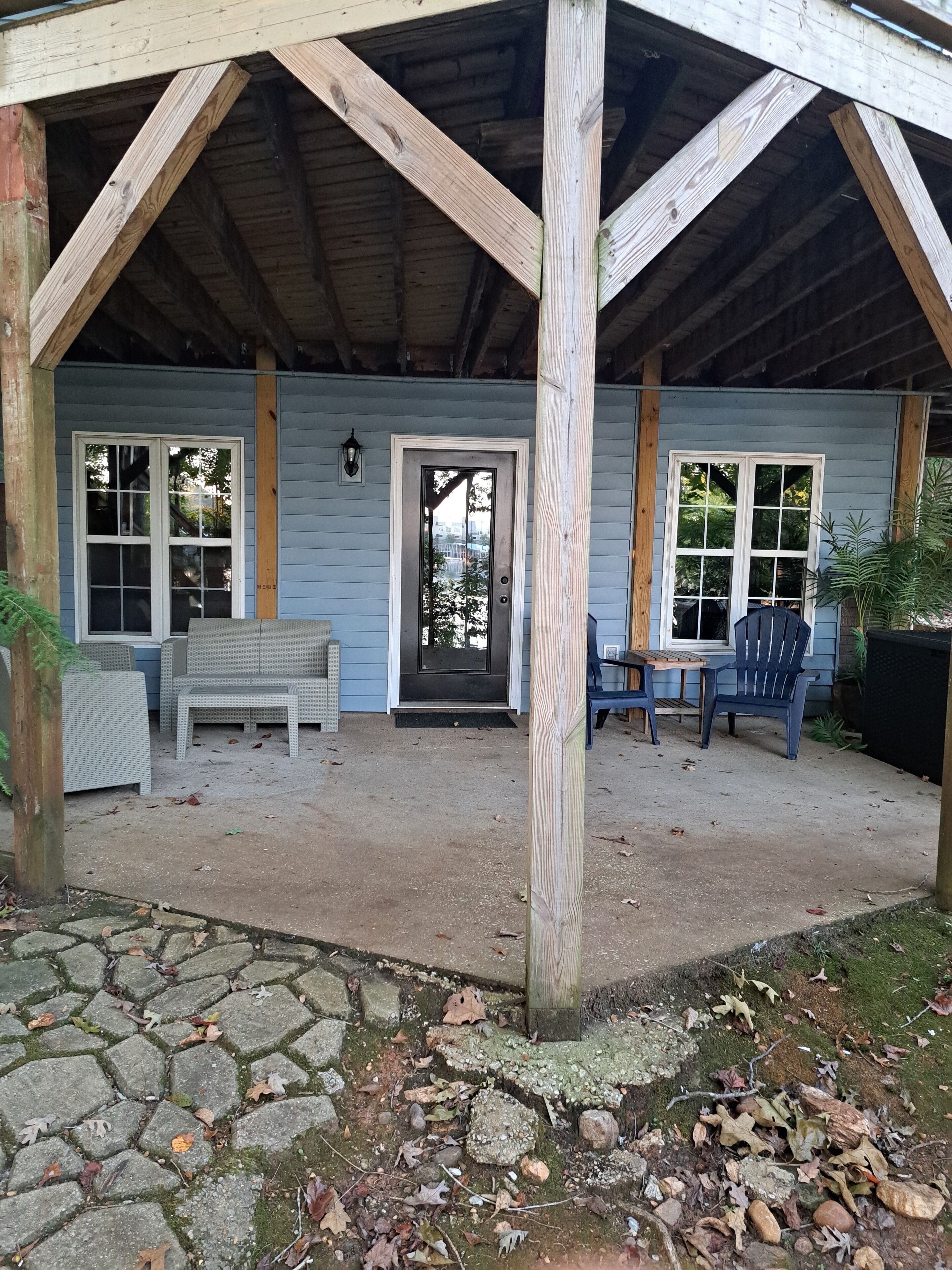 A covered outdoor seating area with a stone-paved floor, wooden beams, and a door with a view of the outdoors.