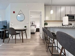 A modern kitchen with white cabinets, a granite countertop, and a microwave. There is a dining area with a table and chairs, and a small plant on the table.