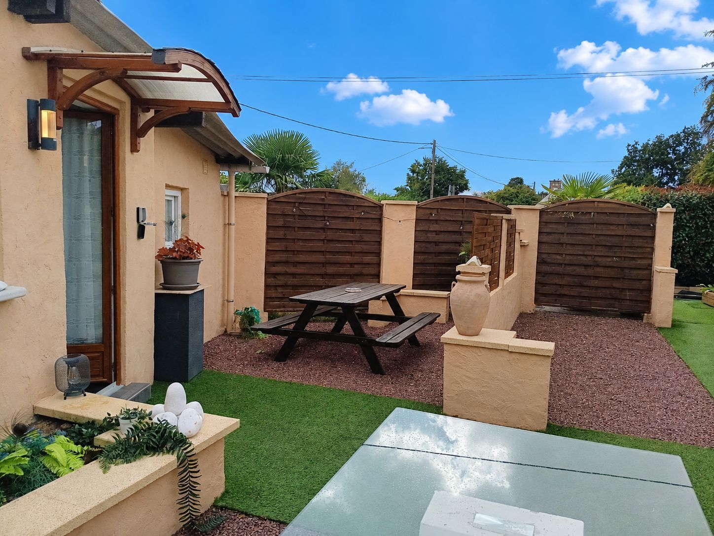 A patio area with a wooden table and benches, surrounded by a low wall and a garden.