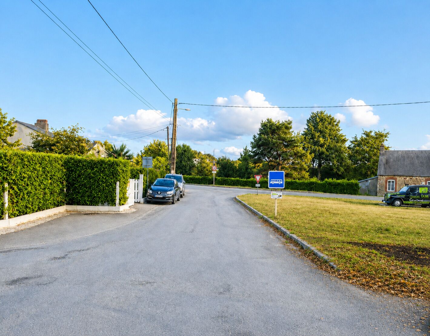 A rural road with parked cars, a bus stop sign, and a house on the right.