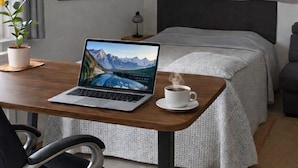 A hotel room with a bed, a desk with a laptop and a cup of coffee, a chair, a potted plant, and a window with curtains.