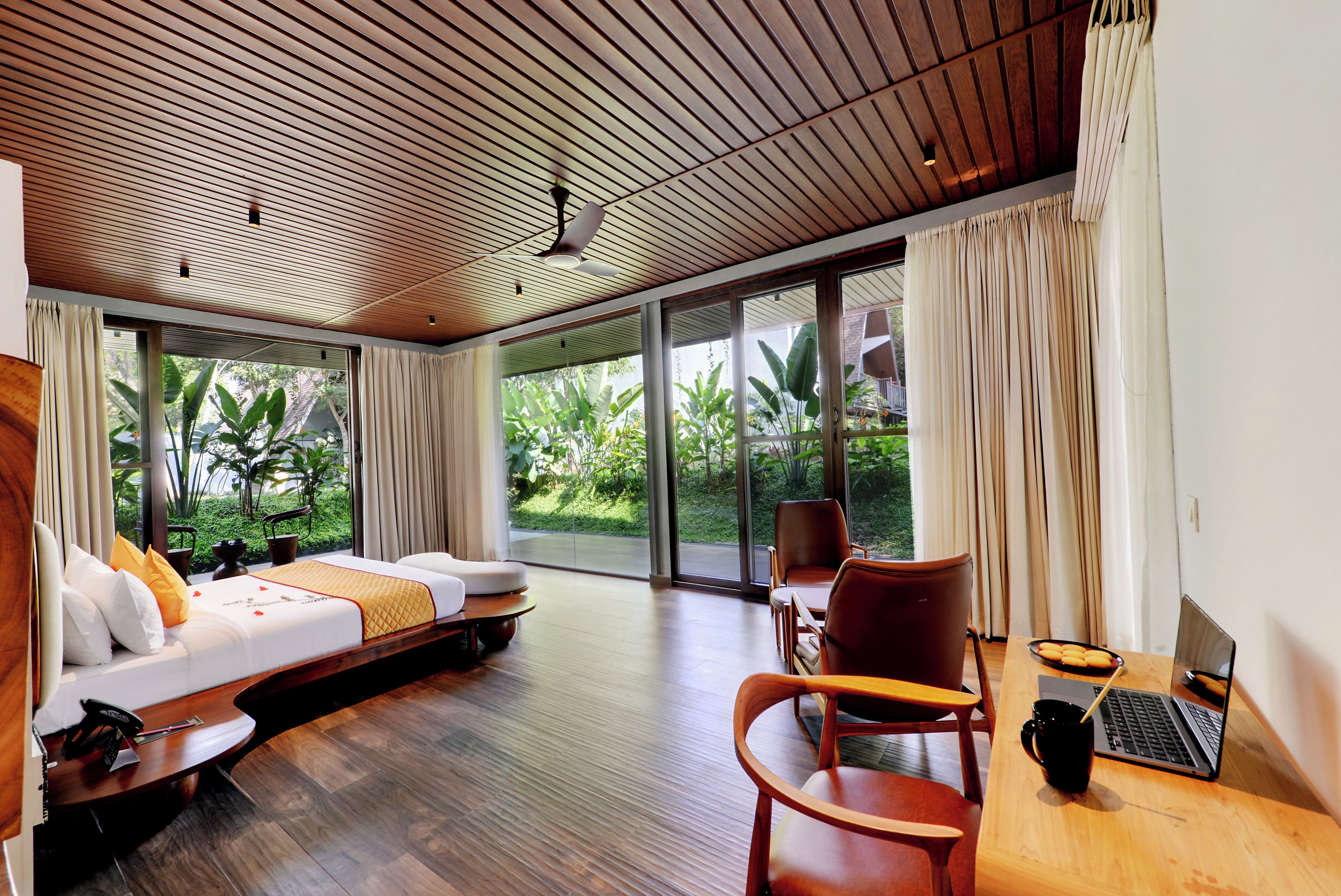 A modern hotel room with a wooden ceiling, large windows, a sofa, a desk with a laptop, and a dining area.