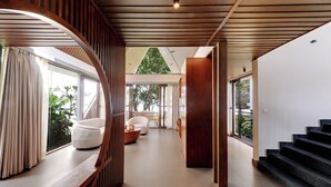 A modern interior with a wooden staircase, a seating area with a round table, and large windows offering a view of greenery. - Sandesh Water Edge Kabini  (Heggadadevankote)
