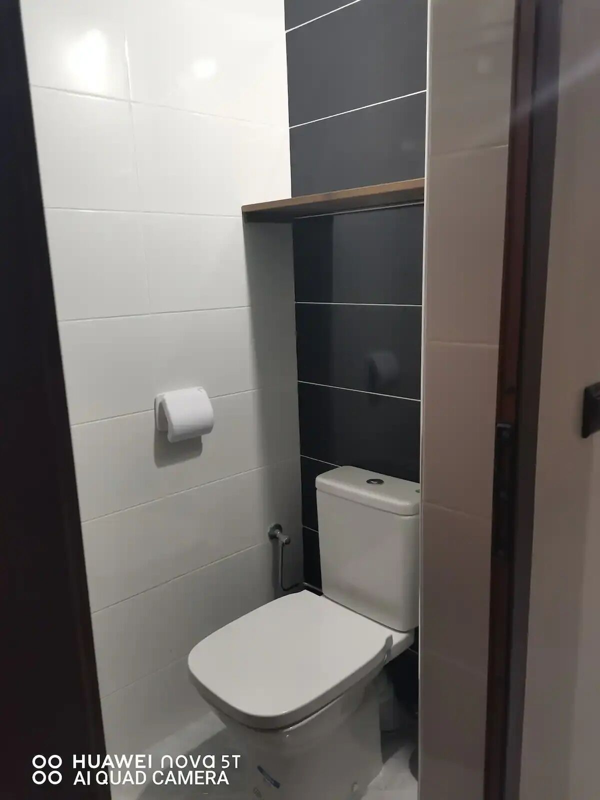 Une petite salle de bain moderne avec des carreaux blancs, des toilettes blanches et une étagère noire au-dessus des toilettes.