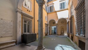 A courtyard with a central column, stone steps, and a plaque on the wall. - Banchi al Patio - Castel Sant Angelo (Rome)