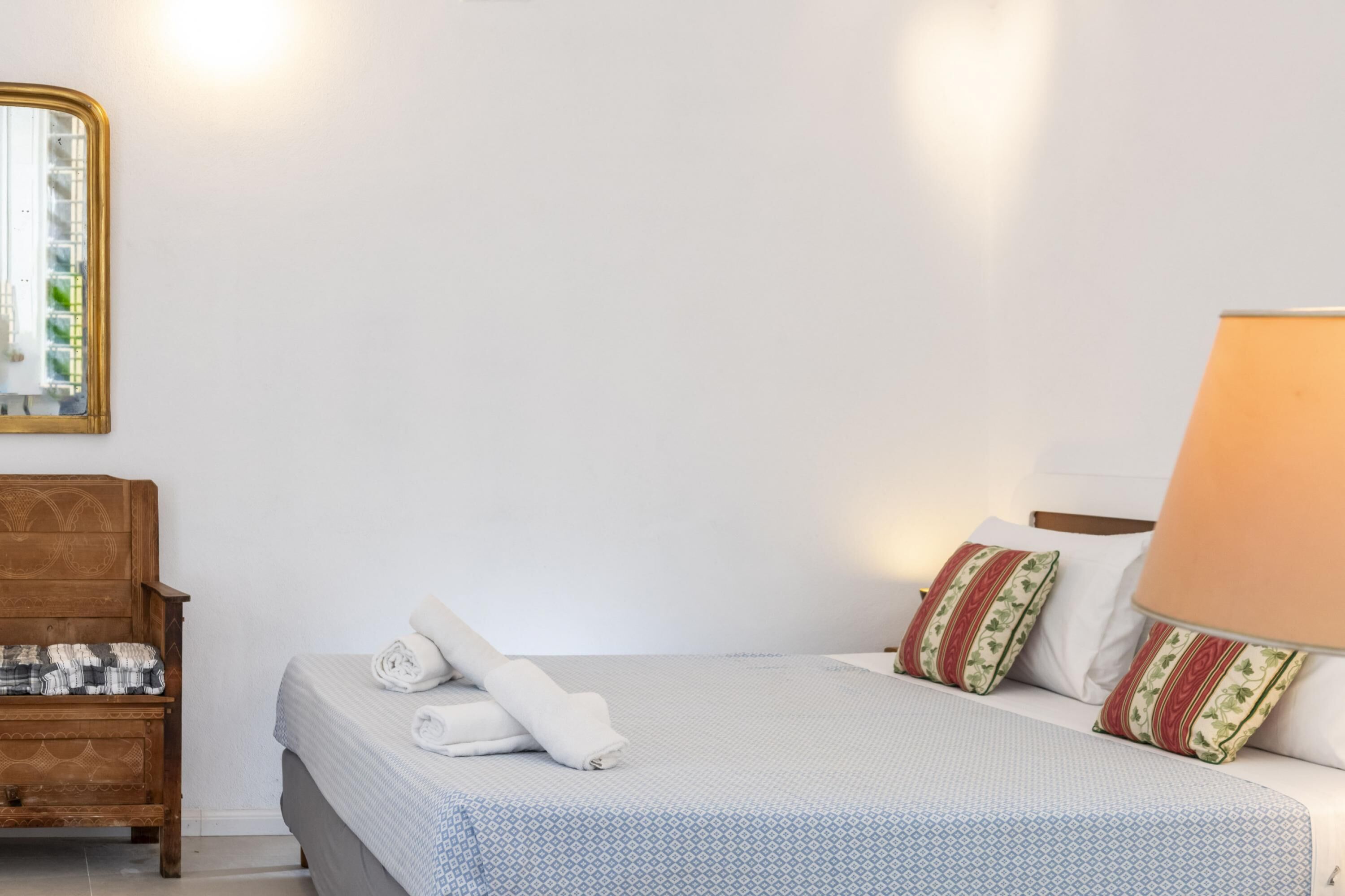 Un lit bien fait, recouvert d’une couvre-lit à motifs, un banc en bois avec une couverture à carreaux, et un miroir fixé au mur.