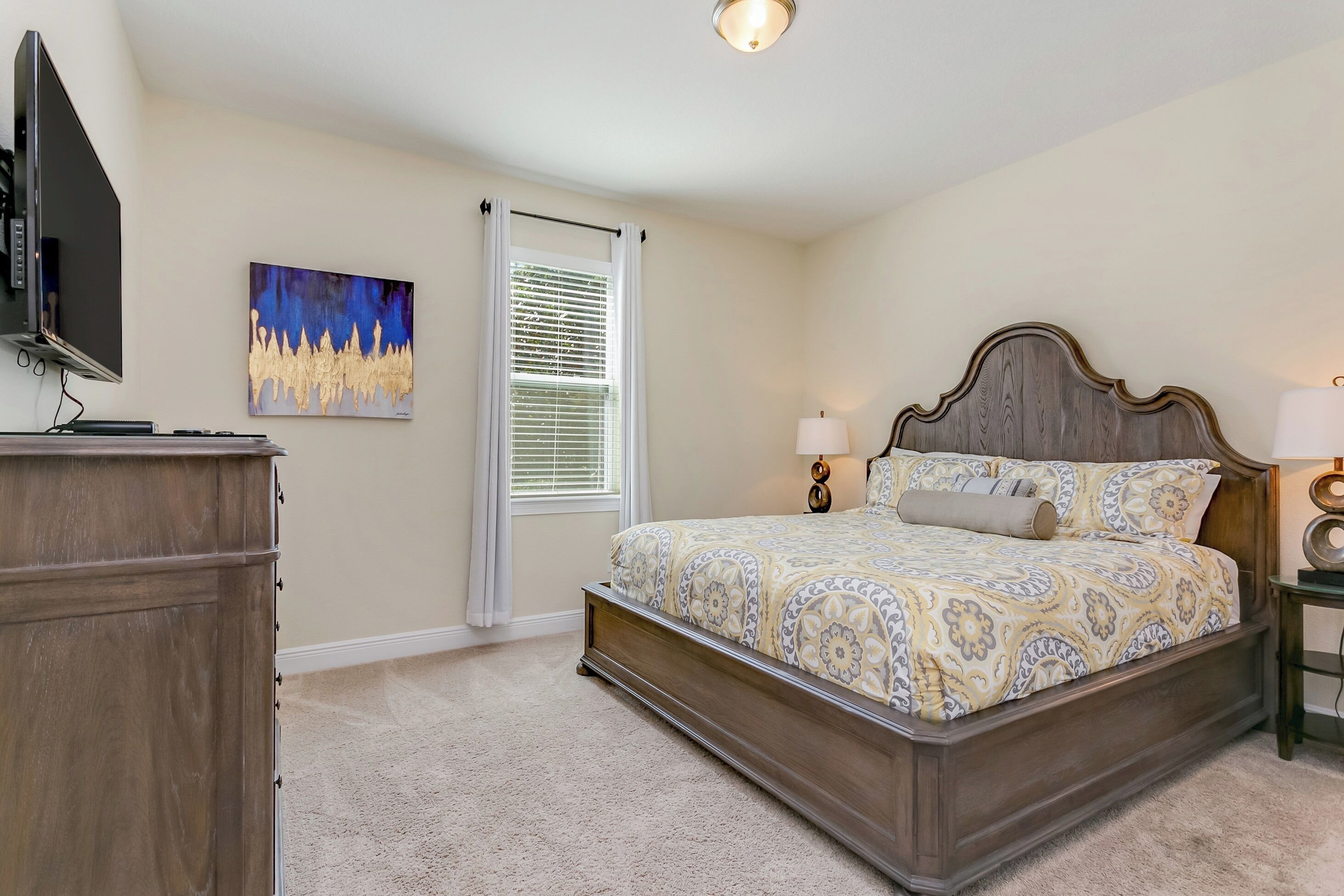 A bedroom with a bed, a TV, a painting, a window with blinds, and a lamp.