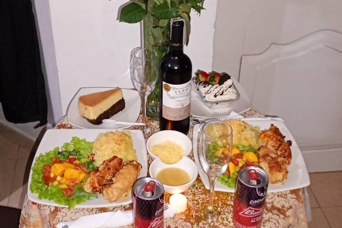 A table set with a bottle of wine, a vase of red roses, and various dishes including a cheesecake, a can of Coca-Cola, and a dessert.
