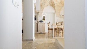 A narrow hallway with a stone ceiling, leading to a dining area with wooden furniture and a white door.