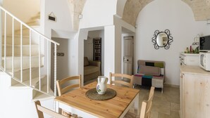 A dining area with a wooden table, chairs, and a vase. There is a staircase with white railings and a view of a living room with a sofa and a television.
